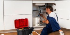 A plumber in blue overalls is fixing a water filter under a kitchen sink. He has a toolbox next to him.